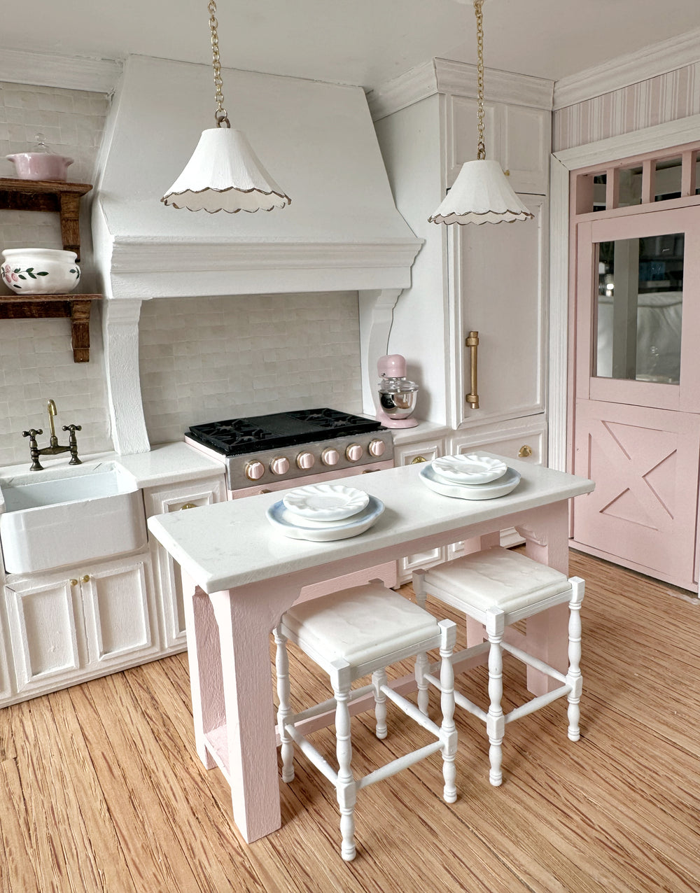 A 1:12 scale kitchen with a pink island, white cabinets and scalloped pendants
