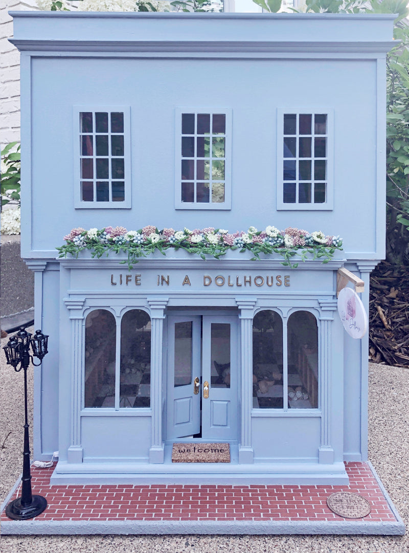 The Life In A Dollhouse miniature shopfront in periwinkle blue with a flower box along the roof and a brick sidewalk